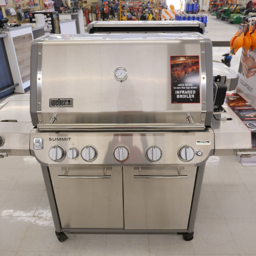 Stainless steel grill with multiple knobs and a thermometer, displayed in a store. Sign reads 