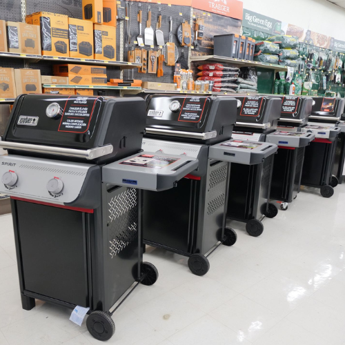 A row of five black and silver gas grills displayed in a store aisle. Shelves above hold grilling accessories, with a bright, organized setup.