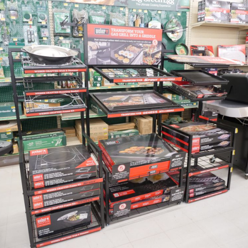 Display of Weber grill accessories in a store. Shelves are stocked with griddles and cookware, promoting a gas grill transformation theme.