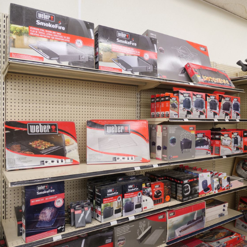 Store shelf displaying Weber grilling products, including SmokeFire pellet grills and accessories in red and black packaging. Brightly lit retail environment.