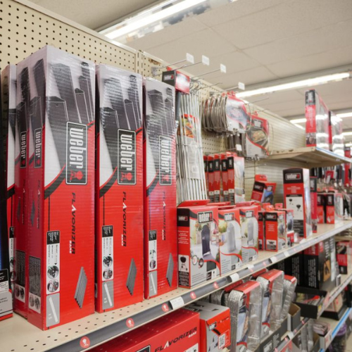 Hardware store aisle with neatly stocked shelves displaying Weber grill parts and accessories in red packaging. Bright lighting creates a tidy, organized feel.