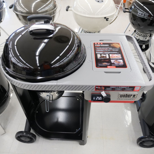 A charcoal grill with a rounded black lid and attached prep surface is displayed in a store setting. Other grills are partially visible in the background.