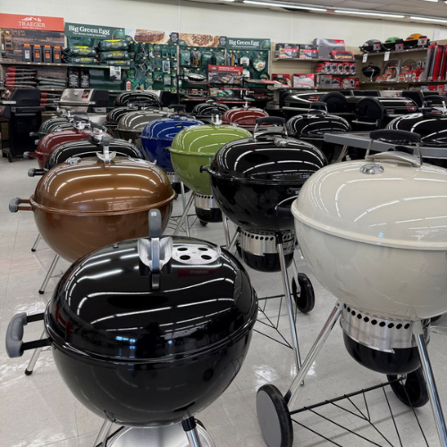 A showroom displaying rows of colorful round charcoal grills. The grills are in black, brown, blue, green, and cream, arranged neatly on a tiled floor.