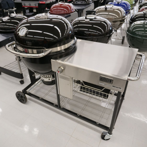 A black and silver outdoor grill with a domed lid and built-in thermometer, placed on a metal cart with shelves, surrounded by similar grills.