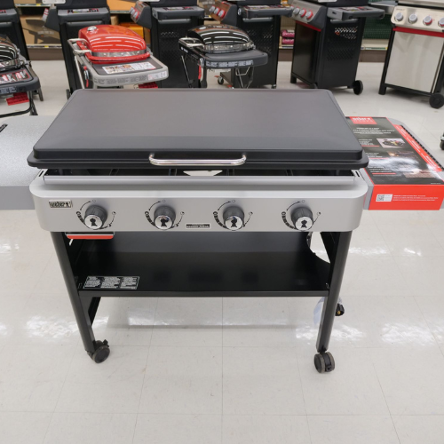 Four-burner gas griddle on display with sleek black and silver design, set on a shiny white floor. Other grills in the background add context.